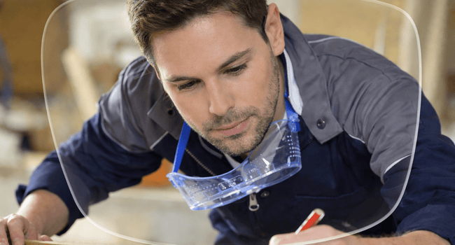 Man in workshop, drawing on wood with pencil. Safety glasses around his neck.