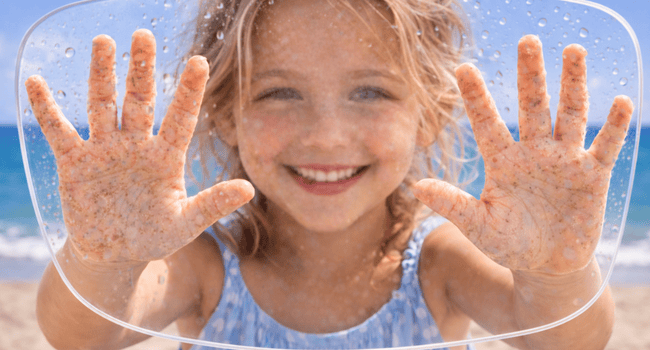 Smiling child with sandy hands, pressed against a clear surface, beach in background.