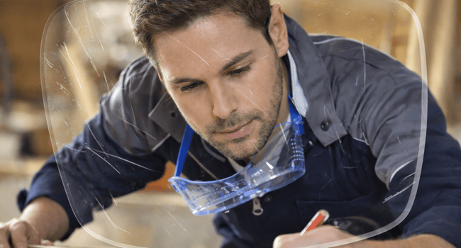 Carpenter in blue overalls, focused, drawing on wood with a pencil. Safety glasses around his neck.