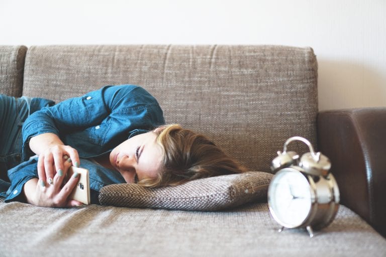 A woman is laying on a couch looking at her phone next to an alarm clock.