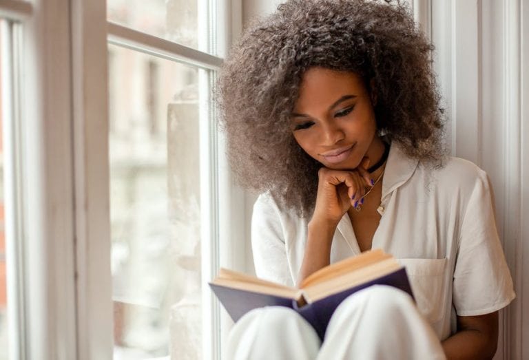 A woman is sitting on a window sill reading a book.