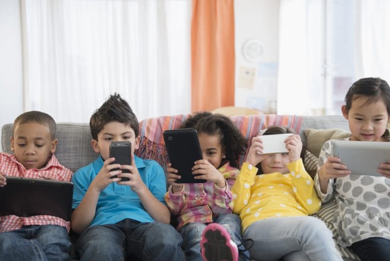 A group of children are sitting on a couch looking at their phones and tablets.