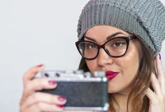 A woman wearing glasses and a hat is taking a selfie with a camera.