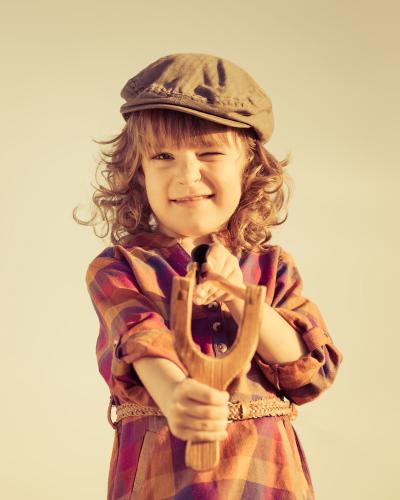 A little girl wearing a hat is holding a wooden slingshot.