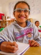 A young girl wearing glasses is sitting at a desk writing in a notebook.