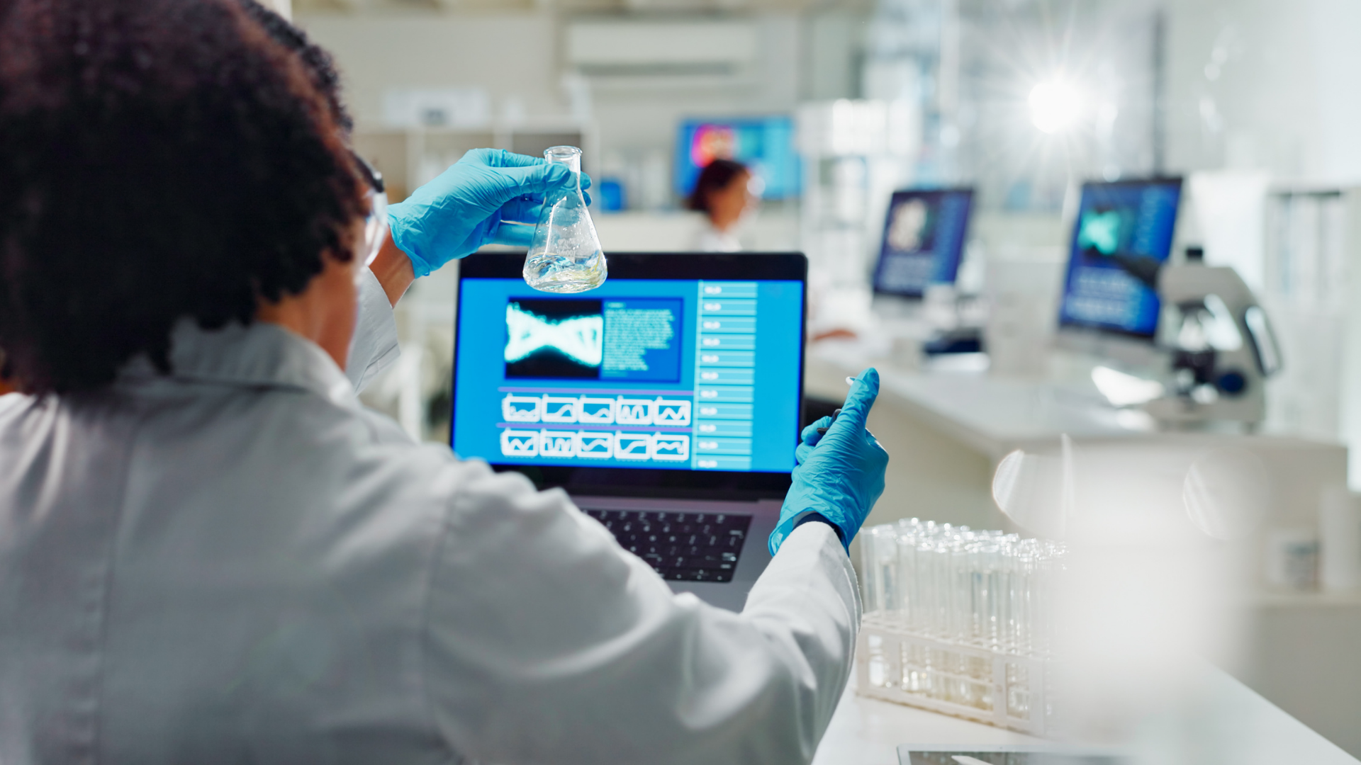 A researcher in a lab coat and blue gloves holds a glass flask while looking at data on a laptop screen.
