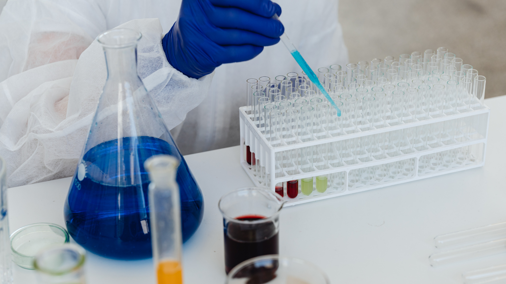 A person in a lab coat and blue gloves uses a pipette to transfer liquid into test tubes on a rack near colorful solutions.