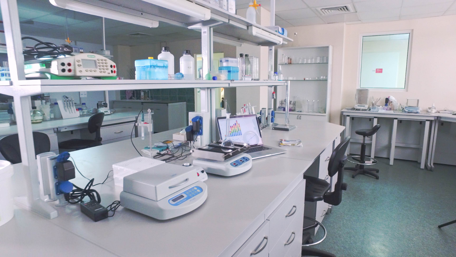 A bright, modern laboratory with white benches, scientific equipment, storage shelves, and stools.