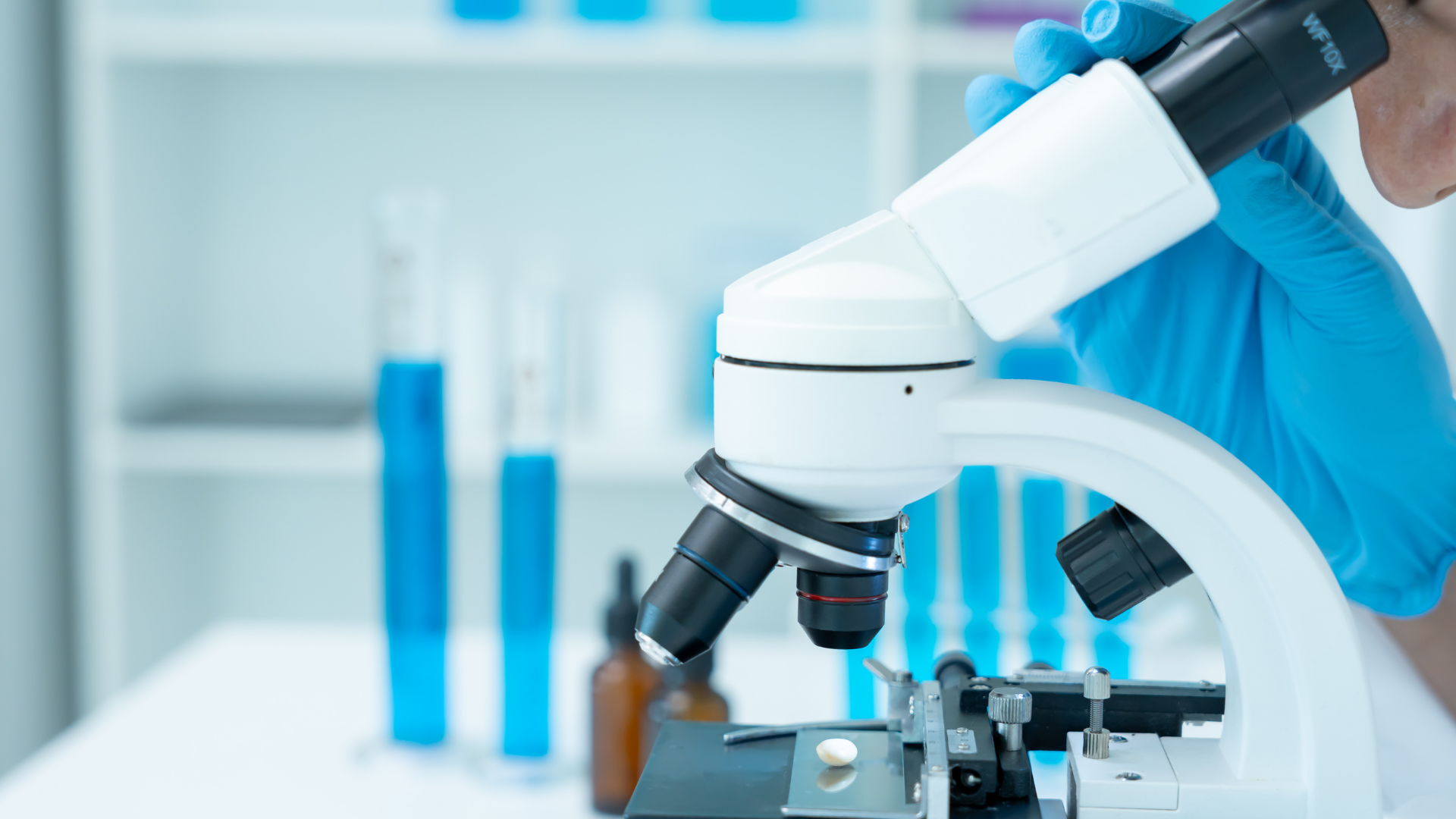A researcher wearing blue gloves adjusts a microscope in a laboratory with blue-filled beakers in the background.