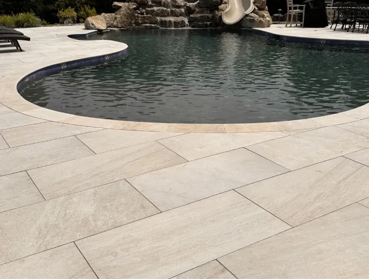 Curved stone patio beside a backyard pool with dark water and rock waterfall feature