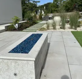 Concrete bench with blue mosaic top beside a tiled patio, lawn, and landscaped garden with steps and fountain grass