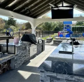 Covered outdoor picnic area with stone tables, black benches, and a grill overlooking a fenced park.