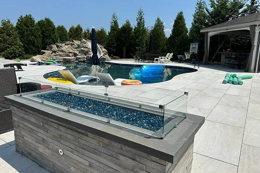 Backyard pool with hot tub, glass fire pit, lounge chairs, and trees under a clear sky