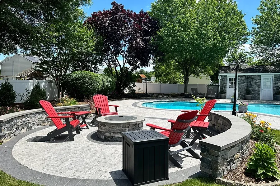 Backyard pool with red chairs around a circular fire pit and black storage bin on a sunny patio