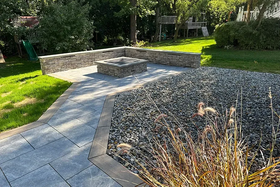 Stone patio with built-in benches beside a gravel garden and green lawn in a sunny backyard