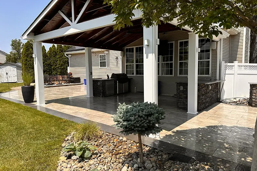 Covered backyard patio with stone flooring, white posts, and outdoor seating beside a lawn and landscaping