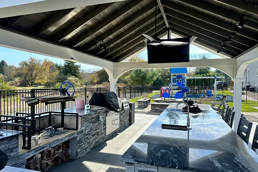 Covered outdoor patio with stone tables, benches, and a TV overlooking a pool and yard