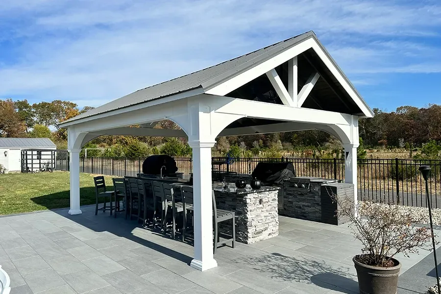 Open-sided pavilion with stone grill, black chairs, and white trim on a paved patio.