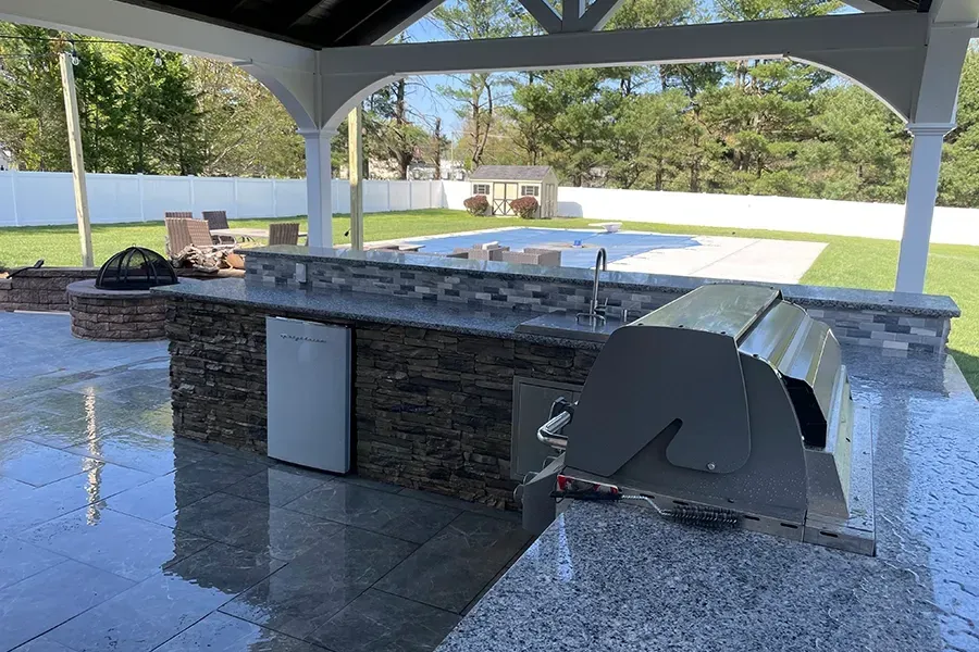 Covered outdoor hot tub with stone bar and grill area beside a pool under a pavilion