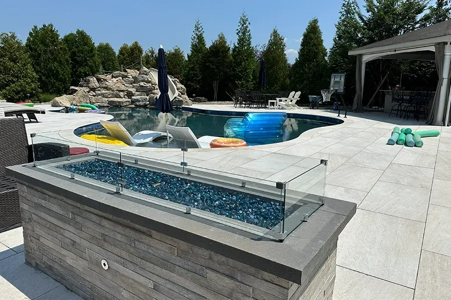 Backyard patio with a pool, hot tub, lounge chairs, and glass fire pit under a sunny sky