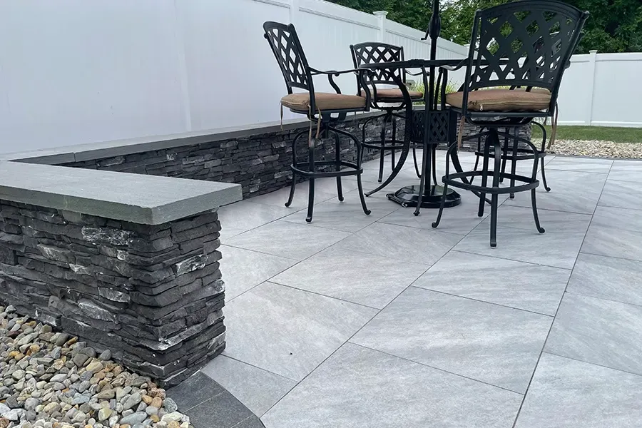 Outdoor patio with black metal table and chairs on light gray stone pavers, beside a stacked-stone bench and white wall