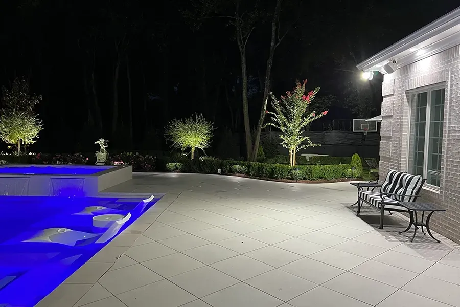 Nighttime patio with blue-lit pool, stone pavers, lounge chairs, and a house exterior.