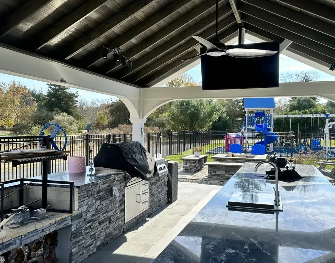 Covered outdoor patio with tables, railings, and a TV overlooking a sunny waterfront park