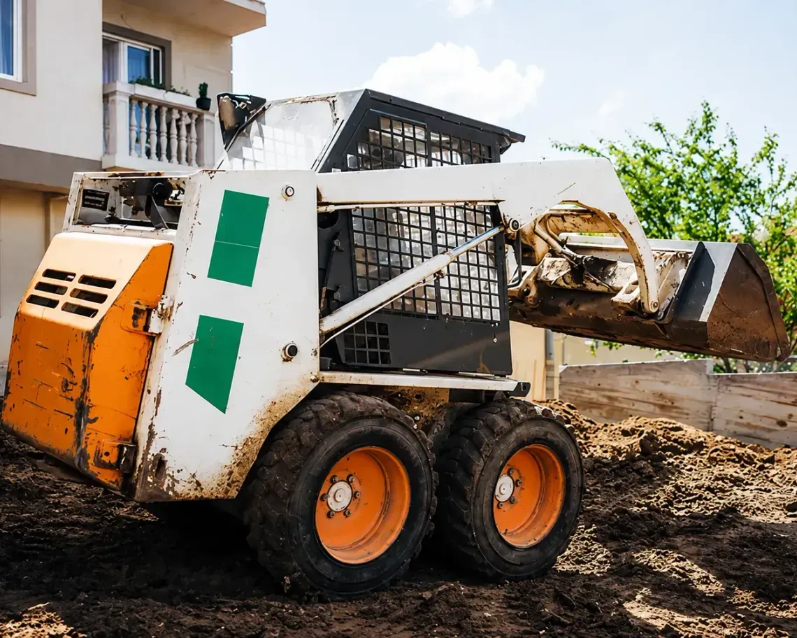 Orange skid steer loader on a muddy construction site beside a house, with bucket raised.