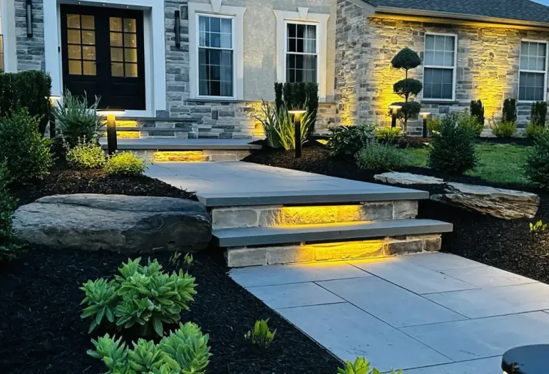 Modern stone house entryway with lit steps, landscaped garden, and dark mulch beds at dusk