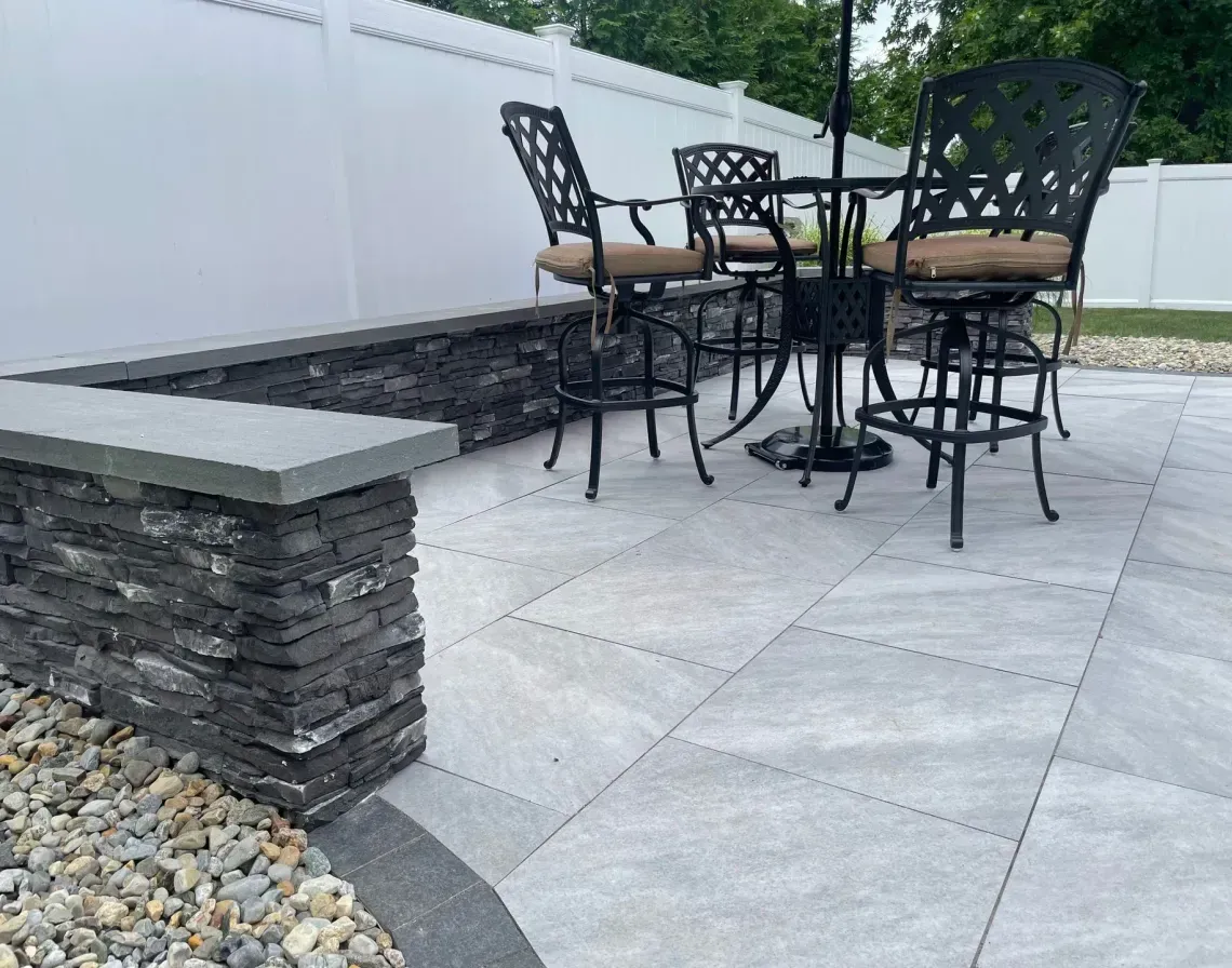 Patio dining set with black chairs and table on a stone-paved backyard next to a raised planter and white fence