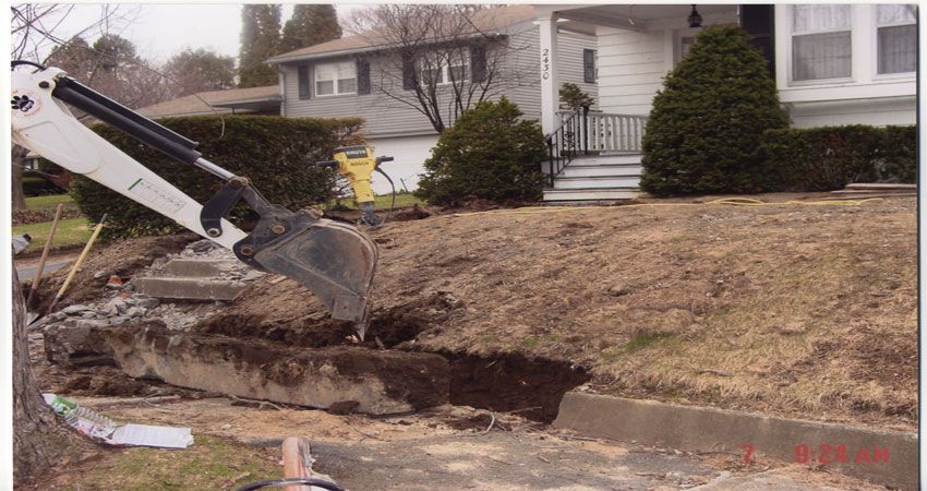 Backhoe excavator digging a trench beside a suburban house and sidewalk