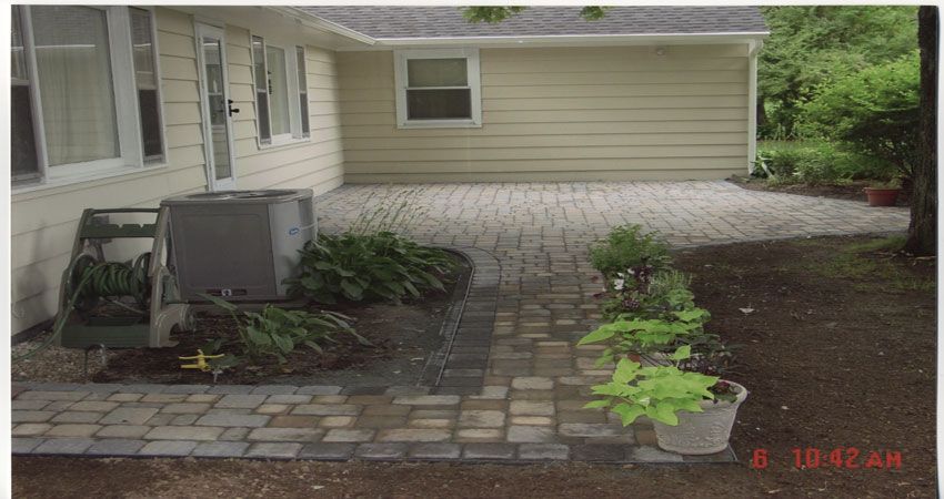 Backyard patio with paver walkway, beige house siding, and small garden plants beside an air conditioner unit