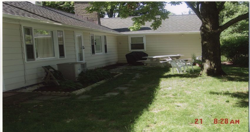 Backyard view of a white house with patio chairs, picnic table, and large trees in shade