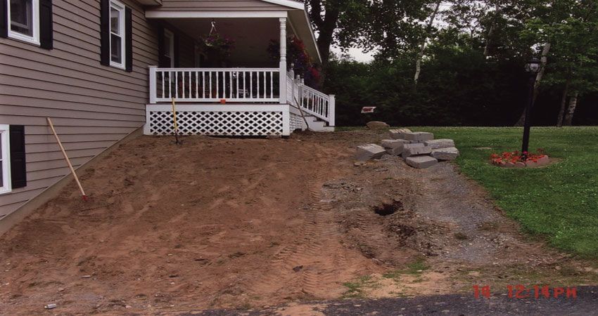 House front yard with exposed dirt slope, retaining wall stones, and a small porch