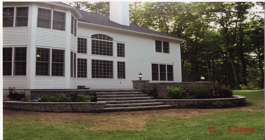 White two-story house with large windows, stone terrace, and wooded yard