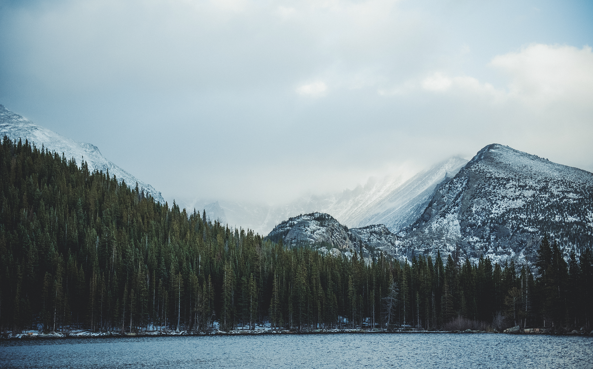 Snow-capped mountains rise behind a dark forest beside a lake under a cloudy sky.