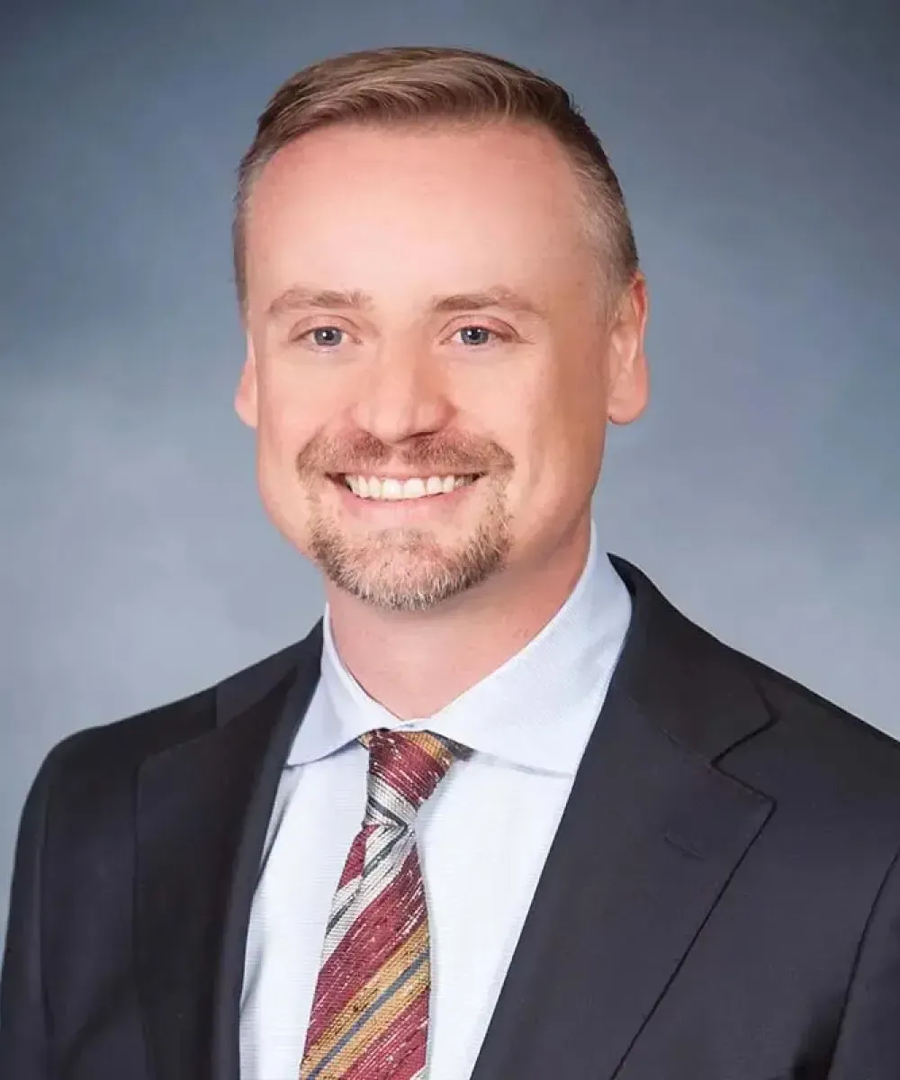Man in suit, smiling, with a patterned tie, against a light blue background.