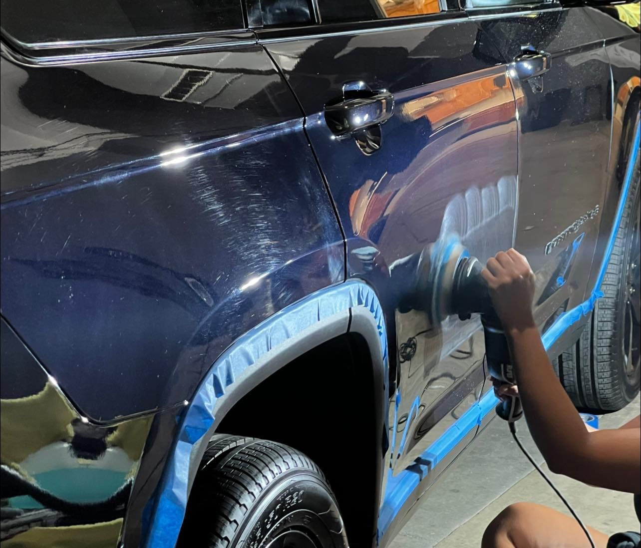 Person polishing a dark blue SUV’s side panel in a garage, with blue masking tape around the wheel arch.