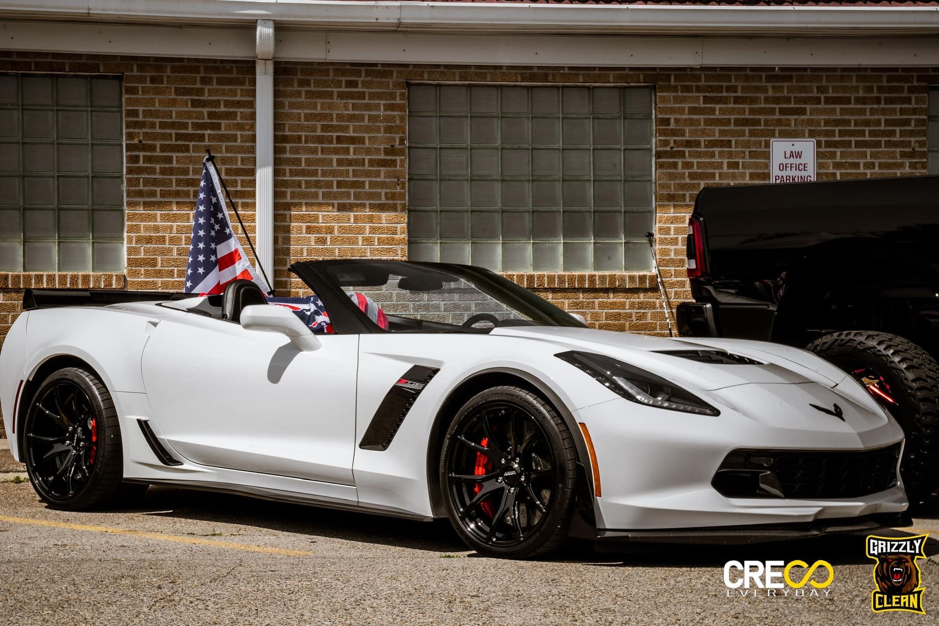 White sports car with black wheels parked outside a brick building, with small flags on top.