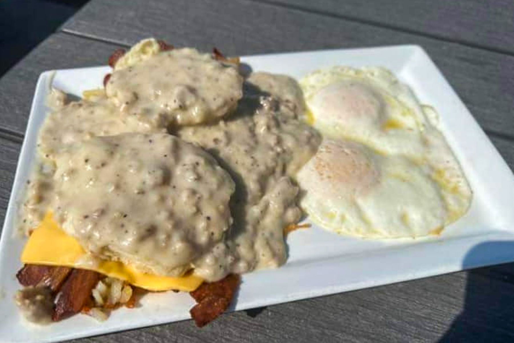 Breakfast plate with biscuit and gravy, cheese, bacon, and two fried eggs on a white tray.