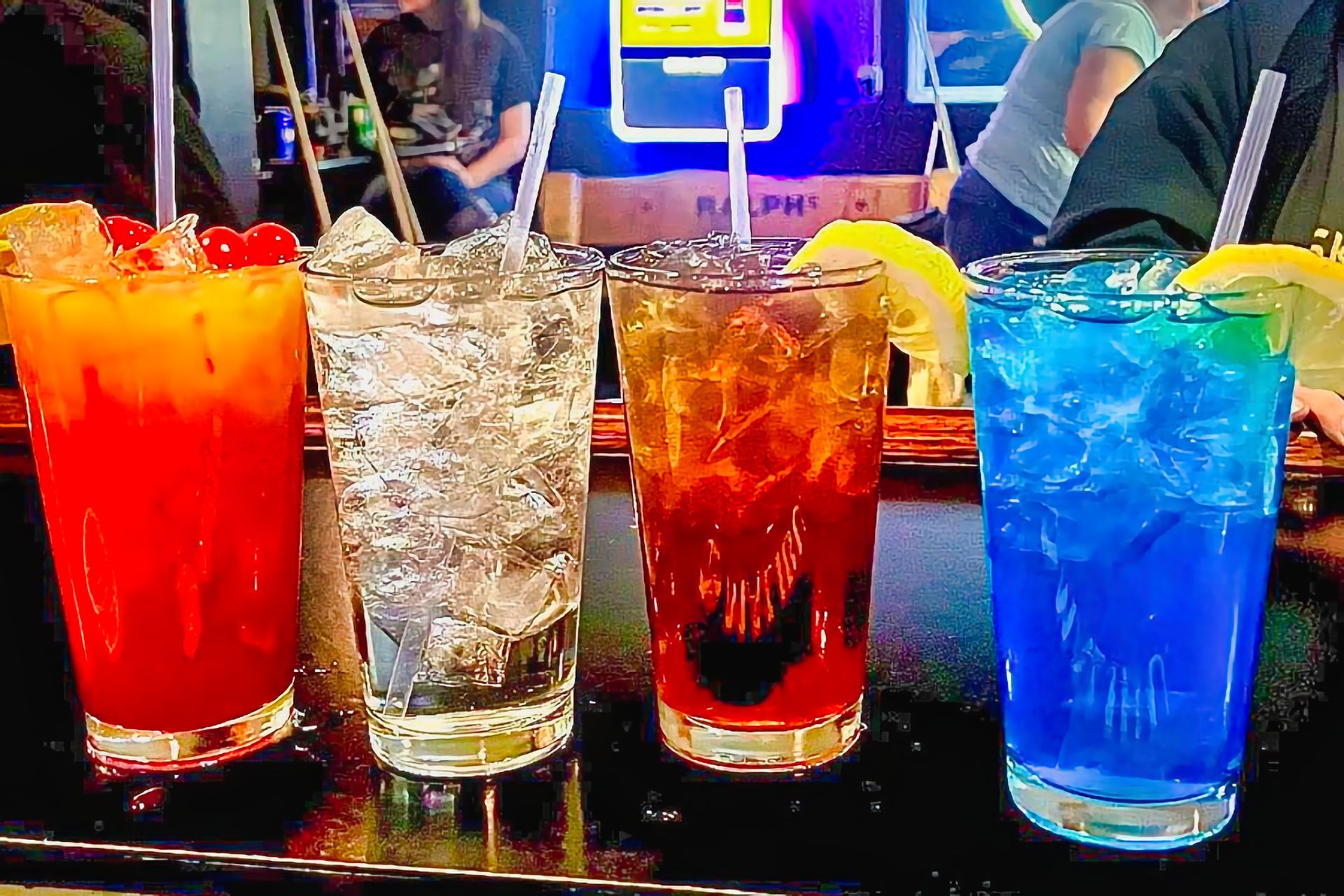 Four colorful cocktails on a bar counter: red, clear, amber, and blue drinks in glasses with ice.