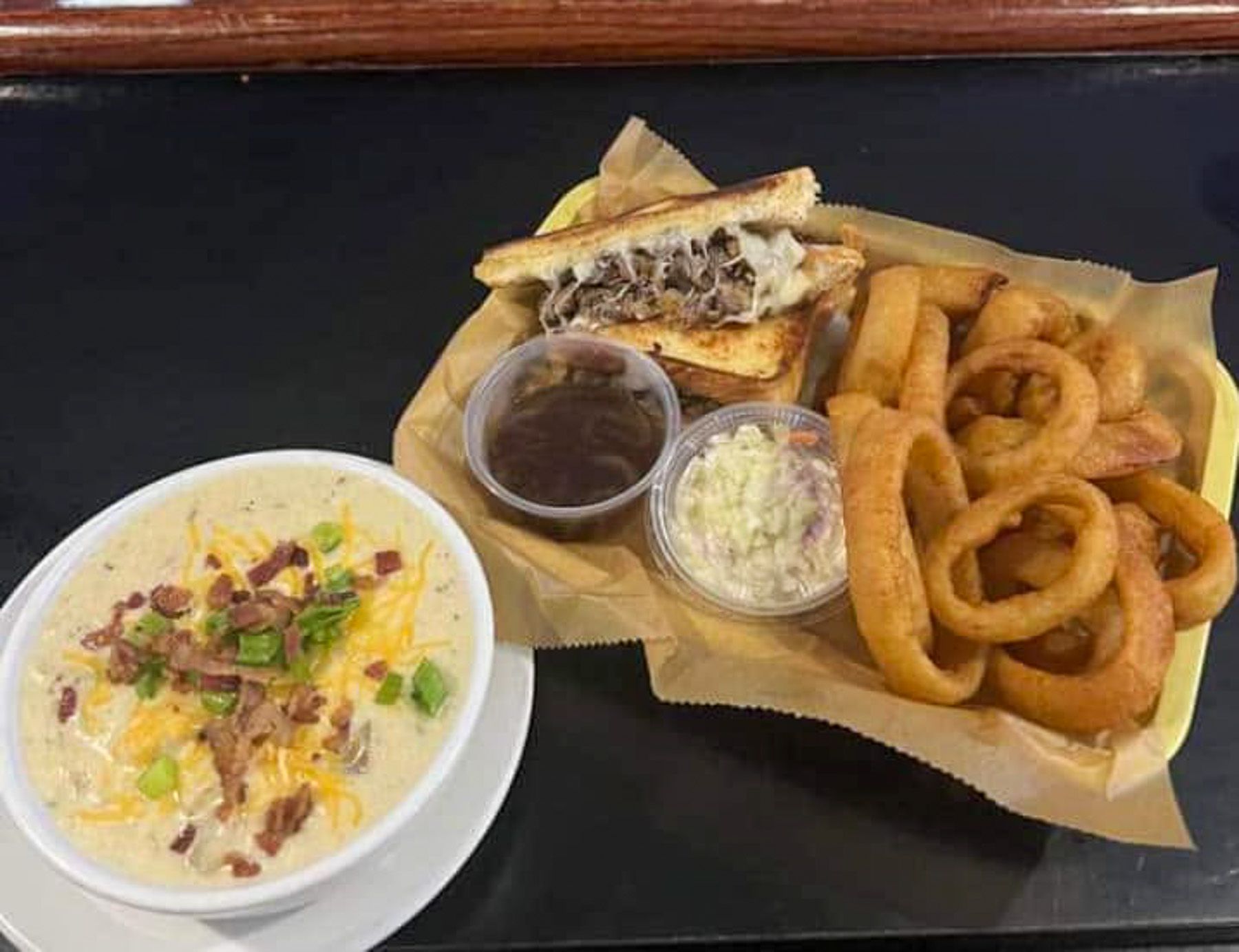 Pulled pork sandwich, onion rings, and coleslaw with BBQ sauce on a tray beside a bowl of loaded soup