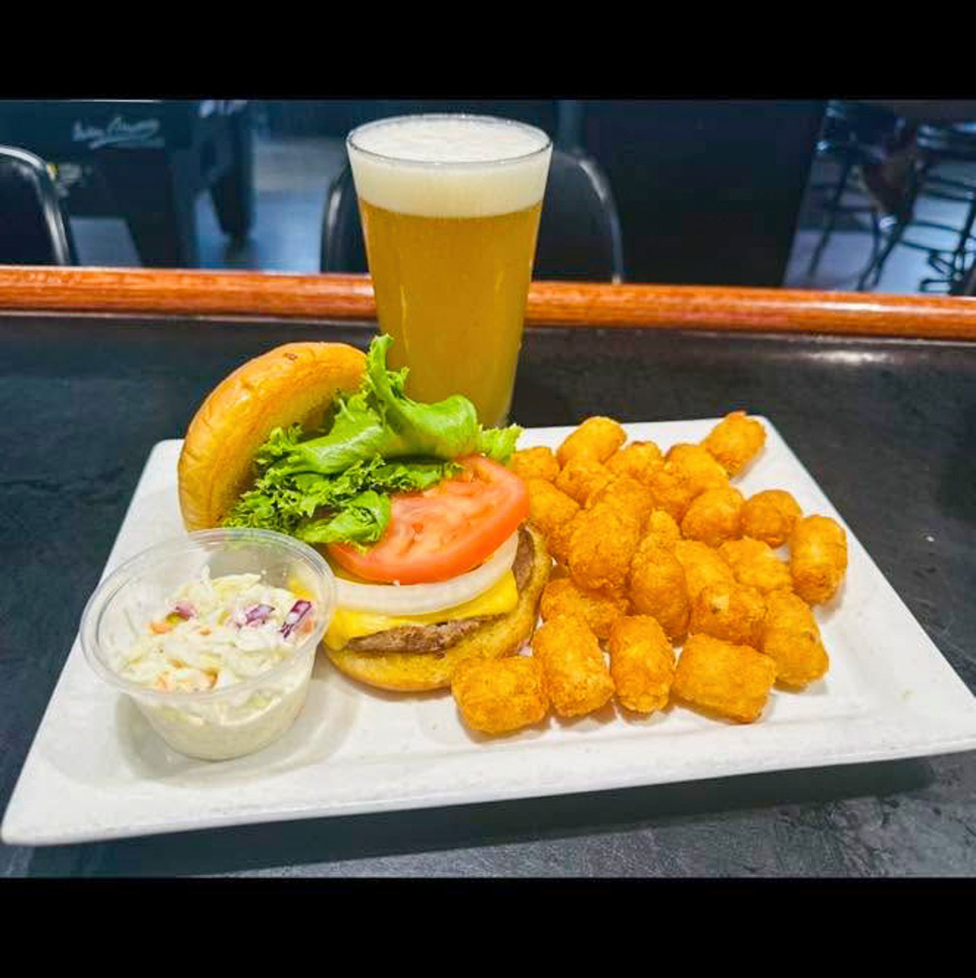 Burger, tater tots, coleslaw, and a beer on a white plate at a bar counter.