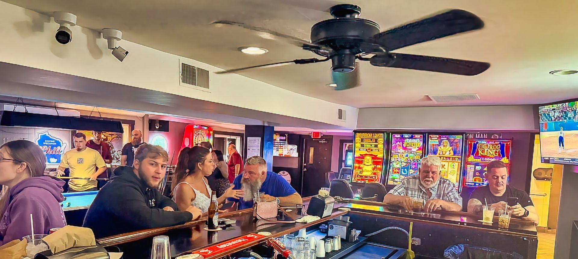 Crowded arcade bar with patrons seated at a counter, neon-lit game machines, and ceiling fans overhead