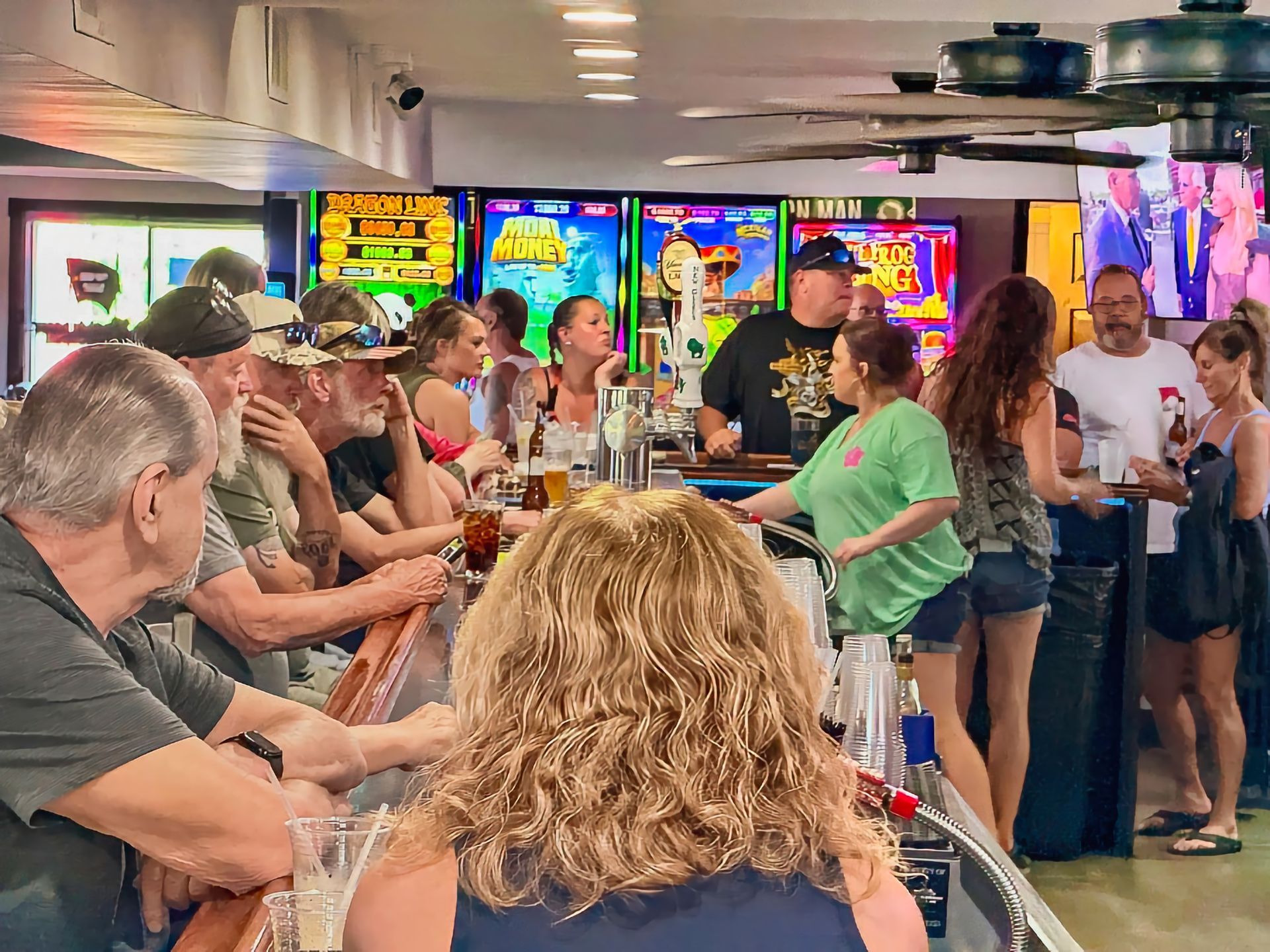 People gathered inside a bar around a counter, chatting under colorful TV screens.