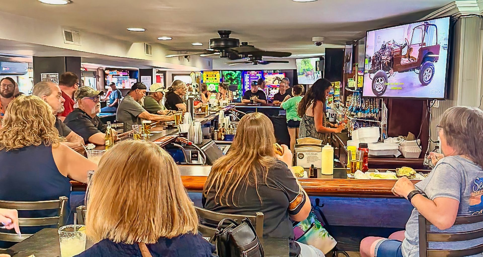 Crowded bar with people seated at tables watching a TV screen and a live performance on stage
