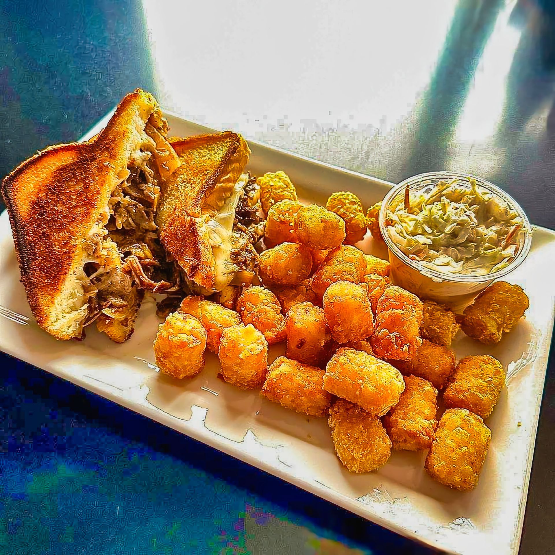 Toast sandwich with tater tots and coleslaw on a white plate by a window