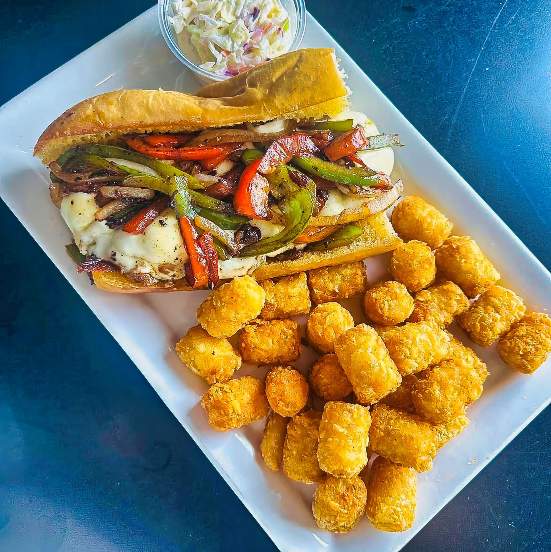 Sandwich, tater tots, and coleslaw on a white plate with a small cup of dipping sauce.