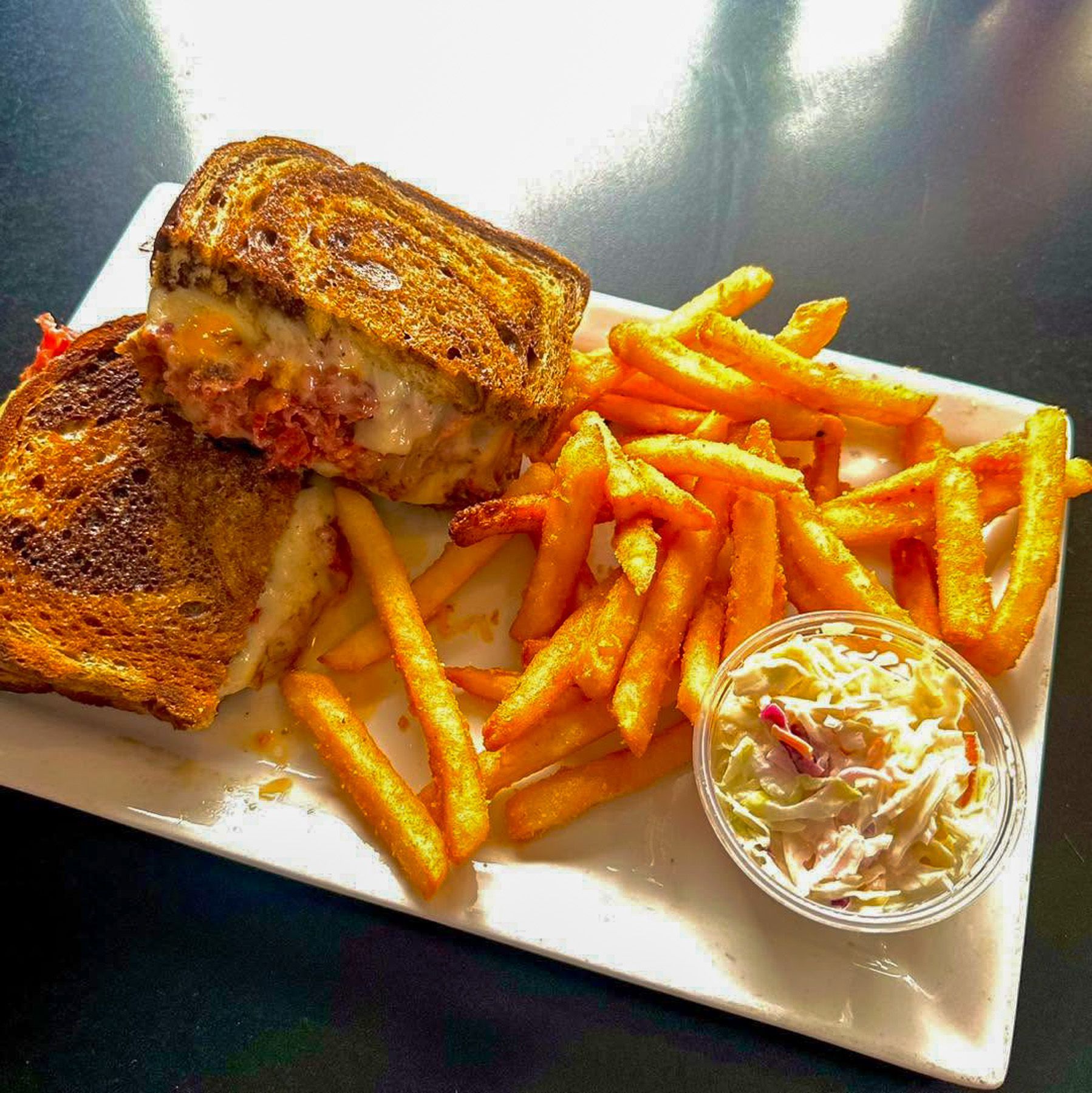 Grilled sandwich with fries and coleslaw on a white plate in bright sunlight