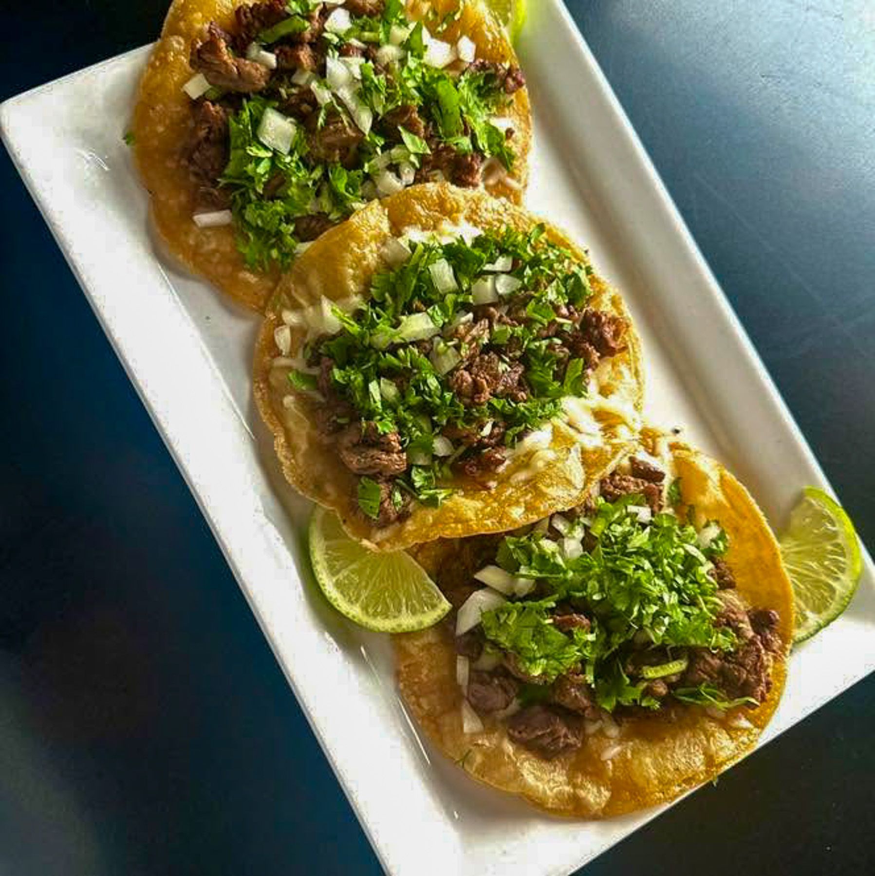 Three tacos topped with chopped cilantro, meat, and lime wedges on a white platter